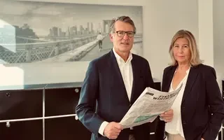 Herr und Frau Tutmann stehen lächelnd in ihrem Büro; Herr Tutmann hält eine Zeitung. Beide tragen formelle Kleidung.